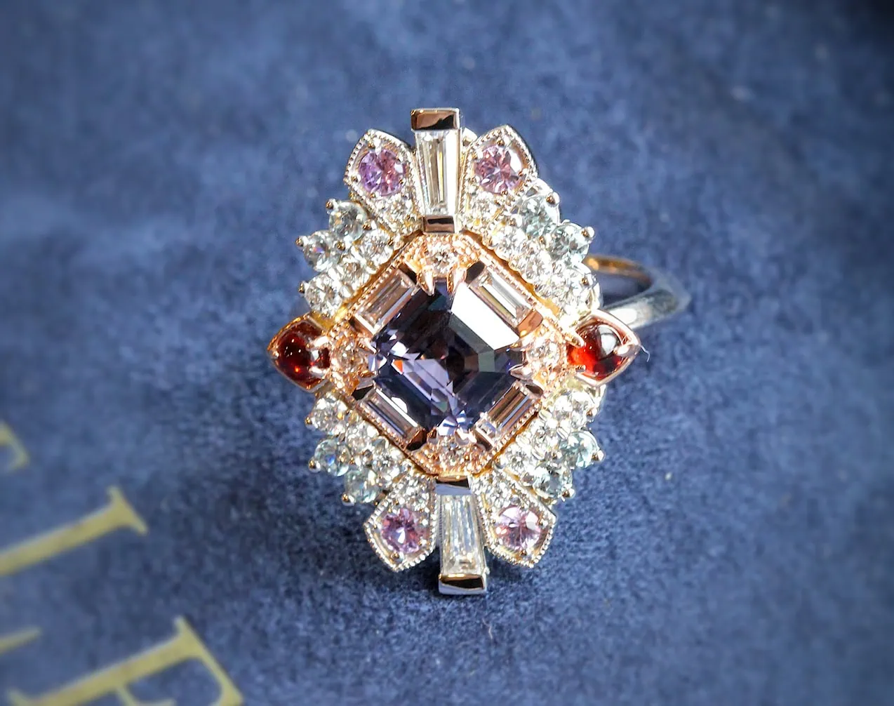 Custom ring with a square-cut center gemstone surrounded by diamonds and colorful accent stones on a blue surface