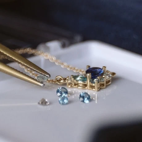 Tweezers holding small blue gemstones beside a pendant necklace setting
