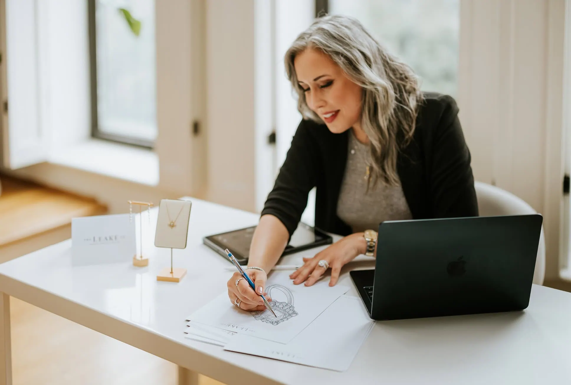 Jewelry designer sketching a ring design at a desk with a laptop and jewelry display